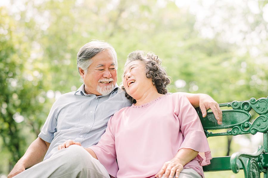 About Our Agency - Senior Couple Laughs Together on a Bench on a Sunny Day in a Park